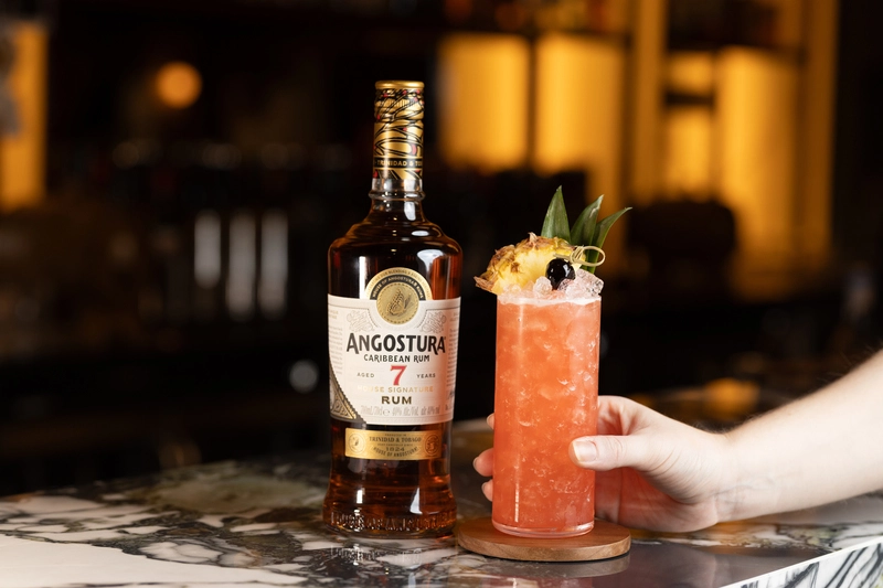 Bottiglia di rum Angostura 7 Years Old House Signature accanto a un cocktail esotico con guarnizione di ananas su bancone in marmo