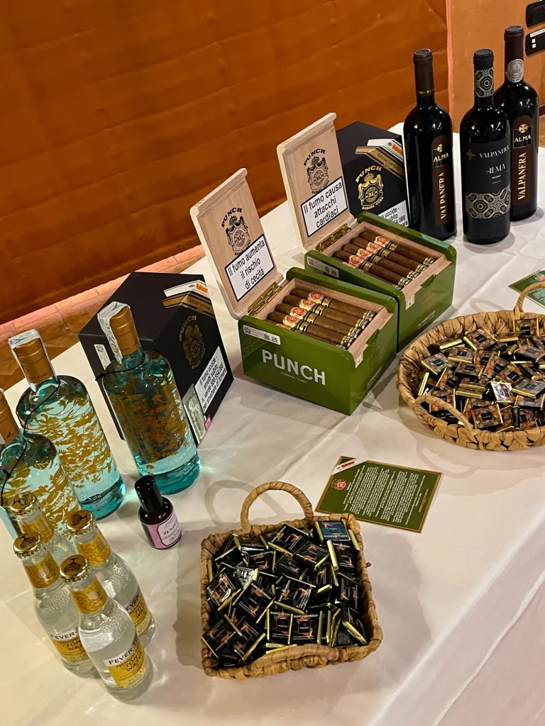 Esposizione sul tavolo delle scatole verdi di sigari Punch, delle bottiglie di vino Alma Valpanera, del gin Silent Pool e del cioccolato Amedei in ceste.