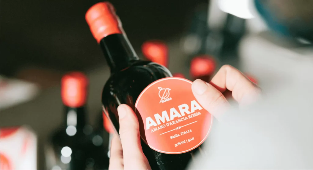 Mani artigiane che applicano l'etichetta rossa sulla bottiglia scura di Amaro Amara