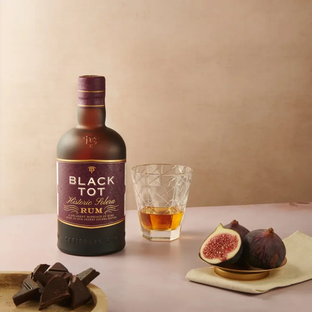 Bottiglia di rum Black Tot Historic Solera sul tavolo accanto a un bicchiere da degustazione, fichi freschi tagliati e pezzi di cioccolato fondente.