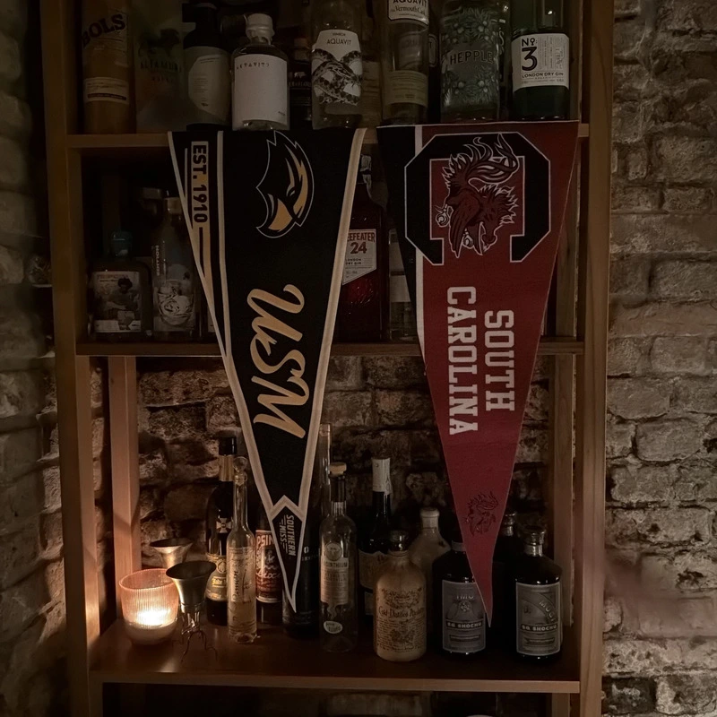 Dettaglio della bottiglieria in legno del 1930 Milano con una selezione di gin e spiriti premium, decorata con gagliardetti universitari americani vintage e mattoni a vista.