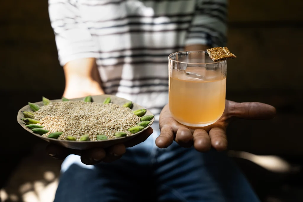 Mani che sostengono un piatto con semi di sesamo e un bicchiere di cocktail artigianale firmato Alquímico.