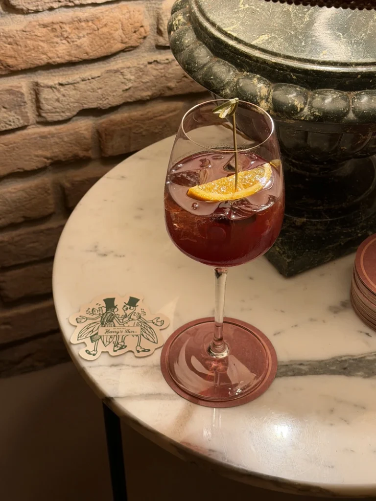 Un cocktail estivo servito in un calice con ghiaccio e guarnizione d'arancia, posizionato accanto all'iconico sticker dell'Harry's Bar di Parigi sul tavolino in marmo del 1930 Milano.