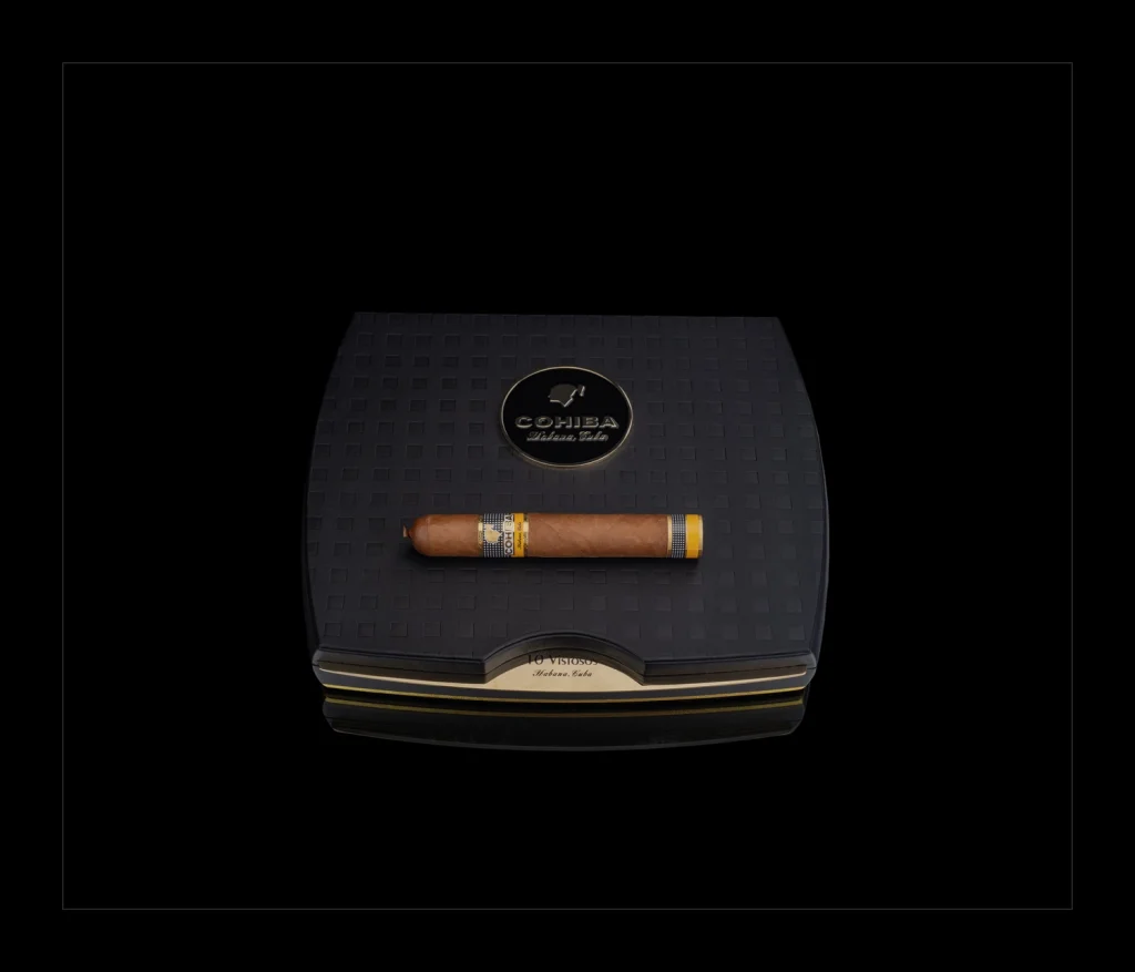 Cohiba Vistosos Travel Humidor chiuso di colore nero con logo dorato e un sigaro Cohiba Vitola Dinoras adagiato sopra.