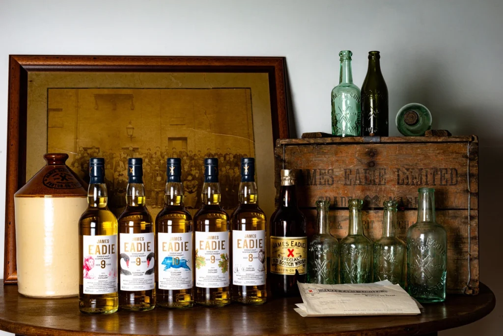 Allineamento di bottiglie di whisky moderne James Eadie Small Batch e antichi cimeli in vetro e ceramica del marchio storico su un tavolo in legno.