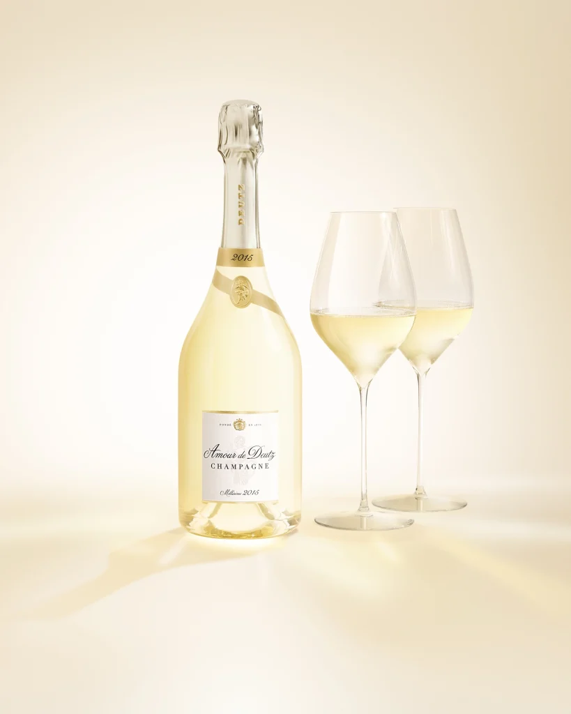 Servizio e degustazione dello Champagne Amour de Deutz 2015 con bottiglia e due calici versati su sfondo luminoso.