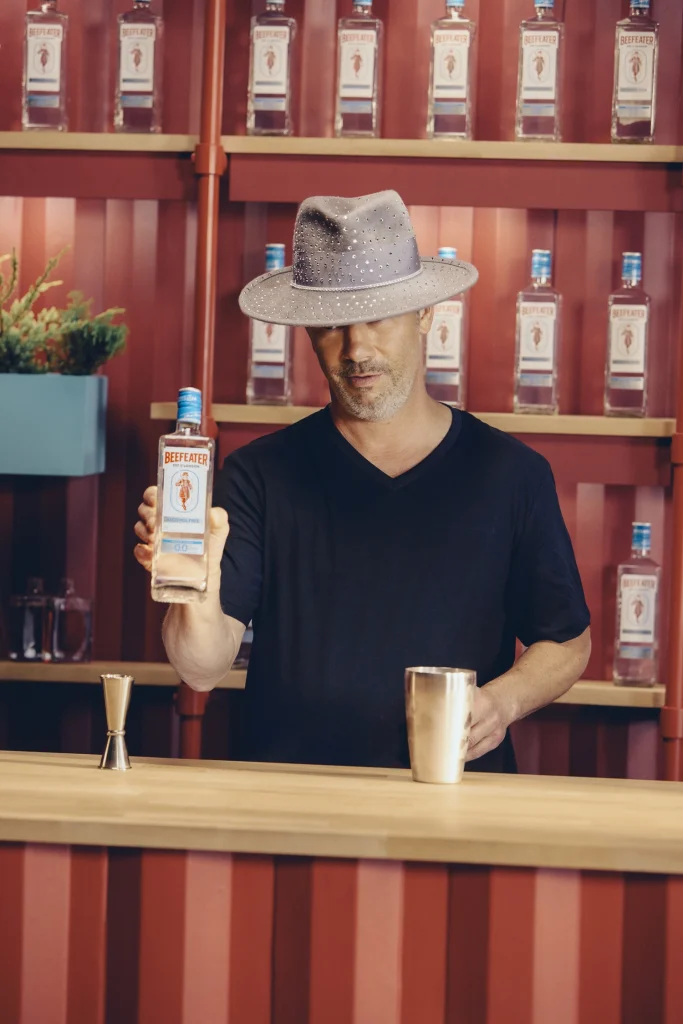 Jay Kay dei Jamiroquai indossa un cappello grigio al banco bar mentre mostra una bottiglia di Beefeater 0.0%.