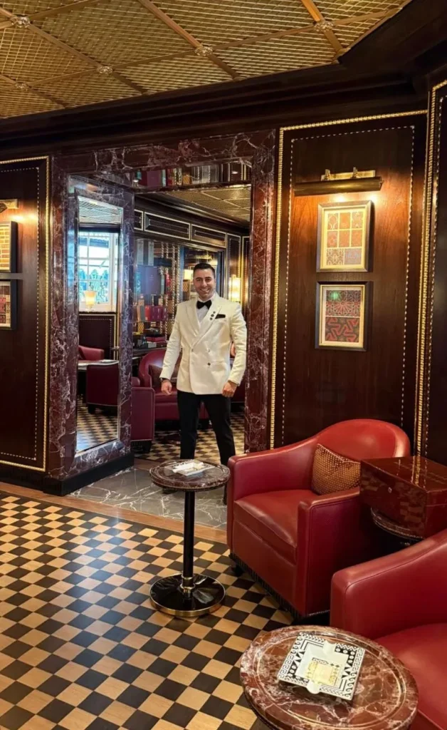 Il Cigar Lounge Manager Luca Ragusini in giacca bianca da smoking sorride all'interno del salotto del fumo lento di The Ritz London, incorniciato da una porta in marmo pregiato.