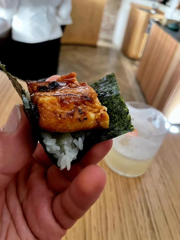 Un nigiri di anguilla su alga nori, con un cocktail d'autore sfocato sullo sfondo al bancone di Akoya.