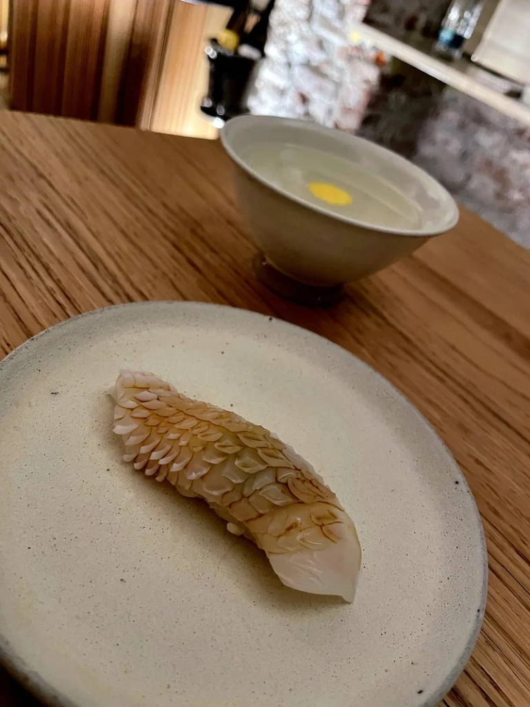 Primo piano di un nigiri di calamaro con un'intagliatura millimetrica a scaglie, accompagnato sullo sfondo da una tazza di brodo dashi limpido.