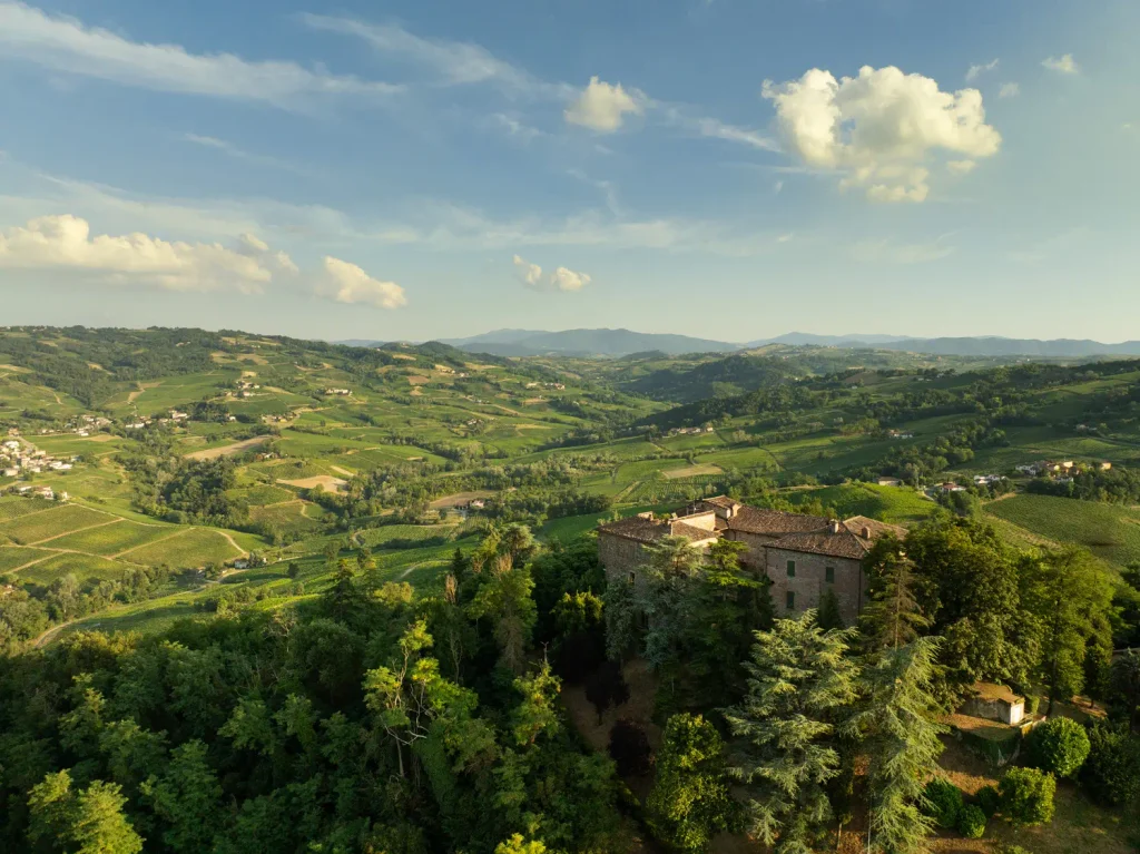 Veduta aerea delle colline dell'Oltrepò Pavese con vigneti di Pinot Nero e un antico casolare in pietra.