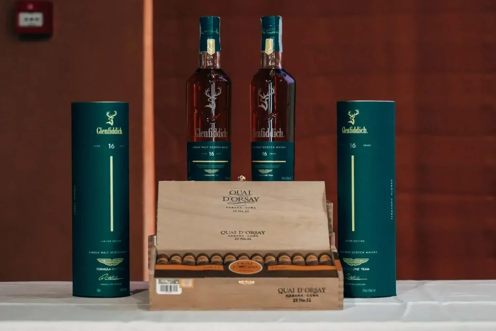Scatola aperta di sigari Quai d'Orsay No.52 disposta accanto alle bottiglie di whisky Glenfiddich 16 Y.O. Aston Martin Limited Edition.