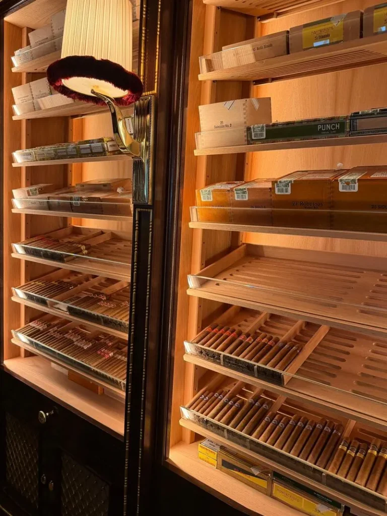 Scatole e file di sigari premium adagiati su scaffali inclinati in legno di cedro umidificato, illuminati a LED all'interno della cabina humidor DeART del Ritz di Londra.