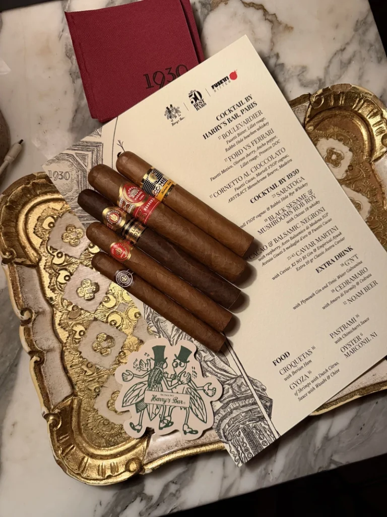 Cinque sigari cubani premium Habanos, tra cui Cohiba e Ramón Allones, adagiati sul menu della drink list dell'evento 1930 Milano e Harry's Bar Paris su un vassoio dorato.