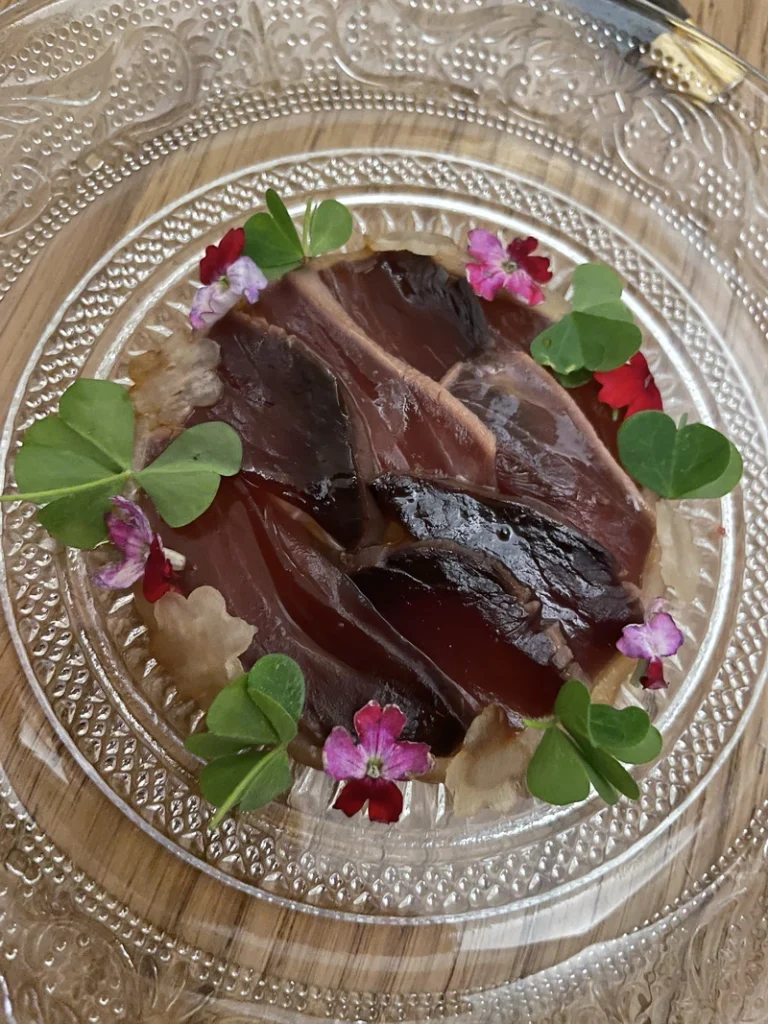 Un piatto di pesce tataki disposto geometricamente su un piatto di vetro, guarnito con fiori eduli e trifoglio.