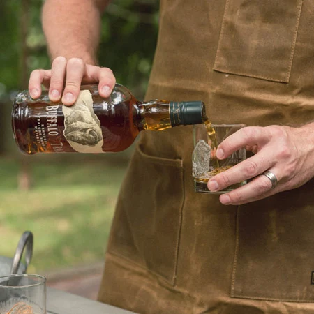 Dettaglio delle mani di una persona con un grembiule marrone che versa del Buffalo Trace Kentucky Straight Bourbon Whiskey da una bottiglia in un bicchiere vuoto. L'immagine si concentra sull'azione di versare.